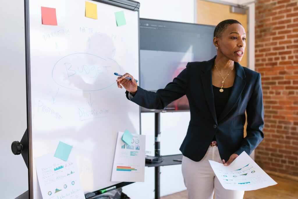 pexels-photo-7414273-7414273 Professional businesswoman explaining budget strategy on a whiteboard during a meeting.